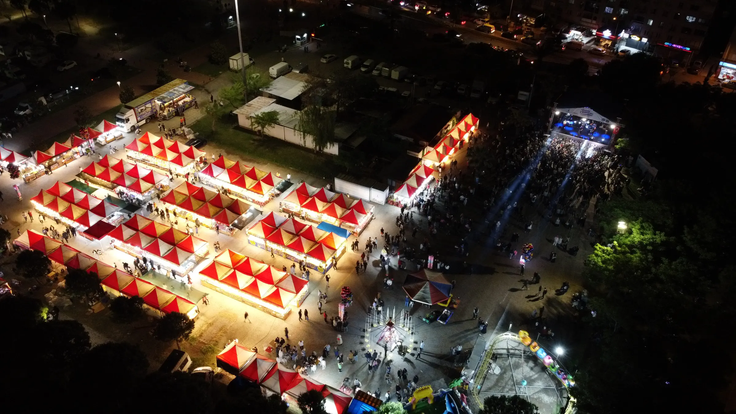A night aerial drone view of a festival ground with lit stalls, a stage and the crowd